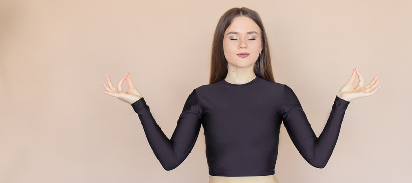 The girl holds her fingers in a "mudra" position and relents with her eyes closed. A young woman on a beige one-ton background.
