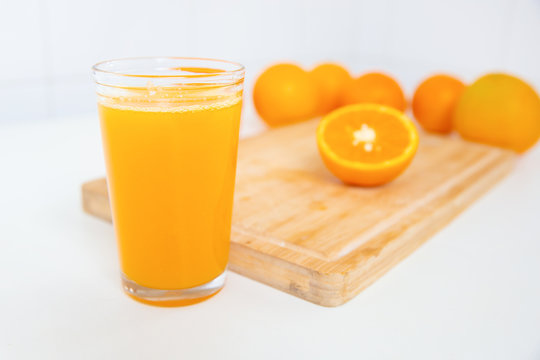 Close-up of glass of orange juice in front of wooden board with two half of orange laying on it. Many juicy fruits out-of-focus on background. Side view. Citrus fruit and healthy food concept