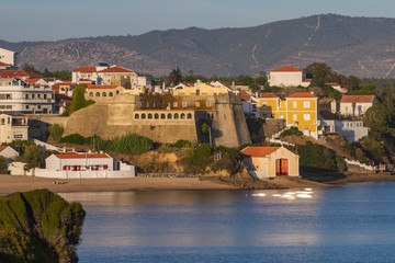 Pueblo pesquero con playa, iluminado por la luz del atardecer con una fortaleza en el centro del...