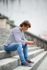 Fototapeta premium Full length side of young man sitting on steps listening to music with earphones and mobile phone