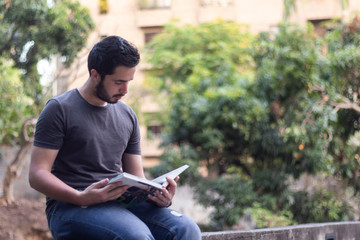 Young man sitting on a wall, holding a book as he reads it, with a garden in the background.