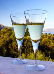 Two Glasses of white wine on seascape background with sun sky mountains and cypress tree landscape reflected in alcohol drink.  An evening cocktail before romantic dinner outdoors on holiday balcony.