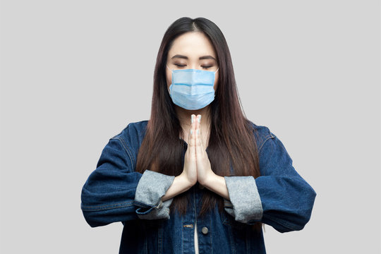 Portrait Of Calm Beautiful Brunette Asian Young Woman With Surgical Medical Mask In Blue Denim Jacket Standing With Palm Hands And Yoga Meditating. Indoor Studio Shot, Isolated On Grey Background.