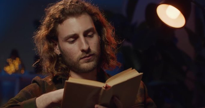 Close up view of inteligent millennial man reading in lamp light while looking sleepy. Handsome man with book turning pages while yawning and feel tired. Concept of leisure.