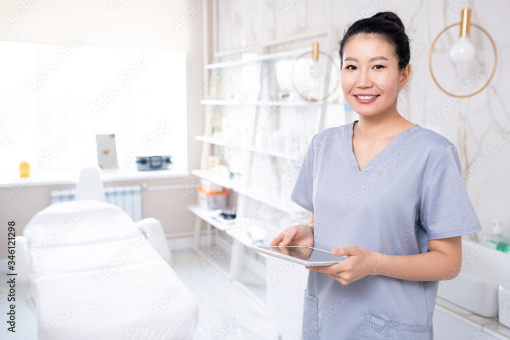 Wall mural Portrait of positive attractive Asian cosmetologist in medical uniform using tablet in clean procedure room