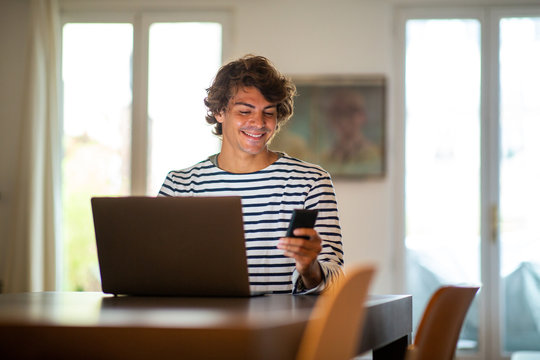 smiling young man sitting at home working with laptop computer and mobile phone