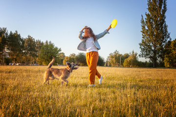 Young beautiful curly girl playing with her dog with a plate of frisbee in summer park