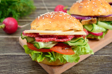Different burgers on a wooden board. Homemade bun with ham or meat or salami, vegetables, herbs. Sandwiches for lunch.