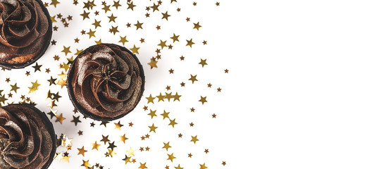 Three delicious chocolate cupcakes with chocolate icing and sprinkled gold sparkles on white background with golden star shaped confetti. Holiday concept. Web banner. Flat lay style. Place for text.