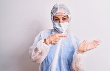 Middle age nurse woman wearing protection coronavirus equipment over white background amazed and smiling to the camera while presenting with hand and pointing with finger.
