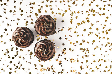 Three delicious chocolate cupcakes with chocolate icing and sprinkled gold sparkles on white background with golden star shaped confetti. Holiday concept. Flat lay style. Place for text.