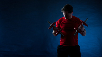 Young non-muscular man doing exercises with dumbbells on dark background