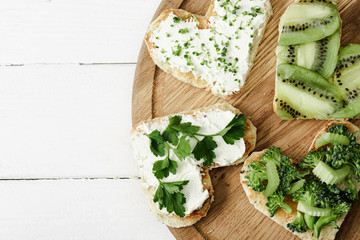 top view of heart shaped canape with creamy cheese, broccoli, microgreen, parsley and kiwi on board on board on white wooden surface