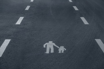 Pedestrian sign painted white on asphalt depicting adult and a child, framed by a dashed lines