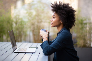 Side of young african american business woman sitting with laptop and cup of coffee - Powered by Adobe