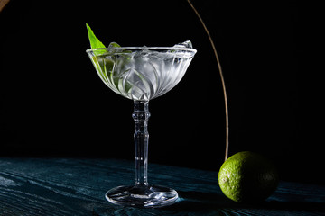 cocktail glass with drink with ice cubes, mint leaf and whole lime on blue wooden surface on black background with geometric lines
