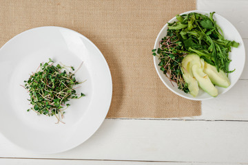 top view of fresh microgreen on plate near bowl of green salad on beige napkin on white wooden surface