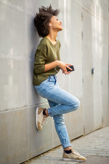 Full body smiling young african woman with mobile phone and earphones leaning against wall
