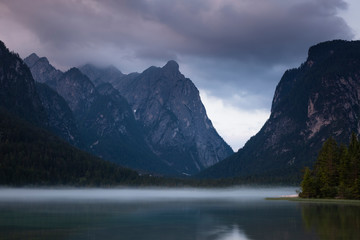 Fototapeta premium Lake Toblach with morning fog in the mountains of the Dolomite Alps during sunset, dramatic clouds in the sky, South Tyrol Italy.