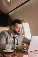concentrated businessman typing on laptop near cup of coffee in private plane