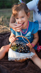 A beautiful and happy girl eats grapes. Mom feeds the baby at a family picnic