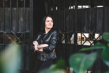 Brunette hipster woman with a positive smile in a leather black jacket posing in the city. Lady model in the Batumi, Georgia. Gates at the background.