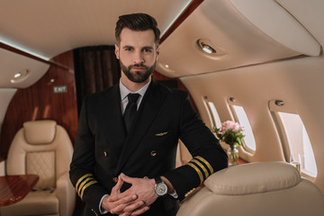 handsome, confident pilot in uniform looking at camera while standing in private jet