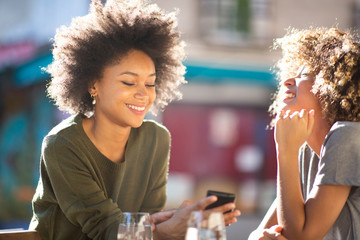 two happy young african american women sitting at outdoor cafe looking at cellphone