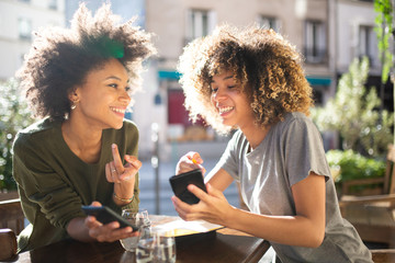 two happy female friends sitting at outdoor cafe with cellphone