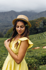 A young girl walk through a beautiful field. Girl in a yellow dress  and a straw hat. Tea plantation, tea garden and green grass.