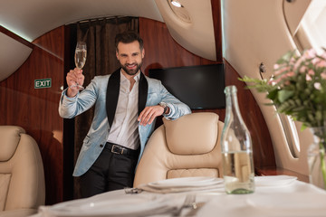selective focus of smiling, elegant man holding glass of champagne and looking at camera in private plane