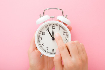 Young woman hands holding white alarm clock. Finger pointing to arrow of twelve o'clock. Light pink table background. Pastel color. Time change concept. Close up. Top down view.