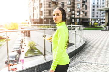 A young girl in trendy sportswear with headphones on the head stands outdoors with water bottle. Street workout