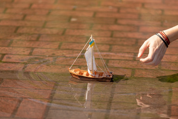 Fototapeta na wymiar Spring. hand starts A wooden toy boat with a sail from medical gauze mask swims along a city puddle. the flag of Ukraine is set on the mast
