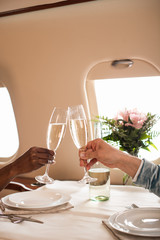partial view of interracial couple clinking champagne glasses at served table in private plane