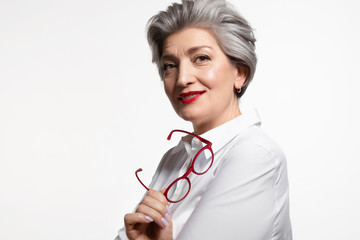 Beautiful mature businesswoman in white shirt with red lips standing with eyewear on white...