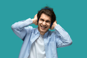 Shock sounds. Portrait of a young beautiful man wearing white t-shirt and blue shirt in headphones covers ears with hands shocked from music