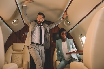 excited businessman screaming and touching ceiling while scared african american businesswoman sitting in private jet