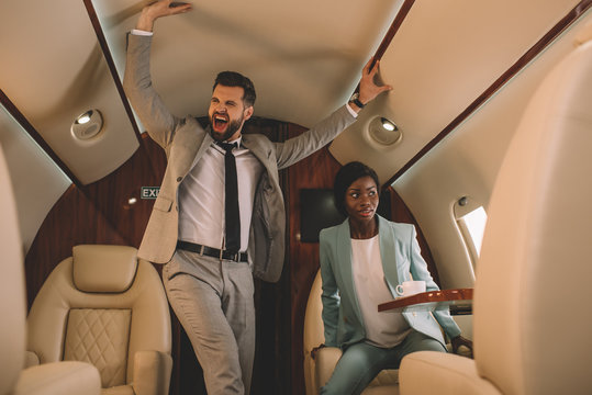 Excited Businessman Screaming And Touching Ceiling While Scared African American Businesswoman Sitting In Private Jet