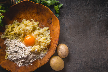 Food background with preparation potato  dough. Potatoes with egg and flour in wooden bowl on dark kitchen table with ingredients. Tasty home cooking.   Top view. Copy space