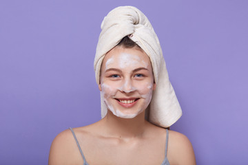 Closeup portrait of smiling happy female posing wrapped in white towel, looking directly at camera, posing isolated over lilac background. Skincare, cosmetology, morning procedures, spa concept.