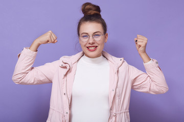 Happy teenage female posing against lilac studio wall clenching fists and celebrating her success,...