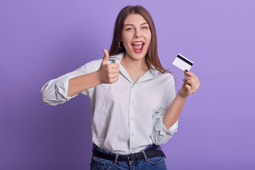 Picture of emotional sweet energetic young woman holding credit card, making gesture, thumb up, showing approval, opening mouth widely, wearing light shirt and jeans. People and money concept.