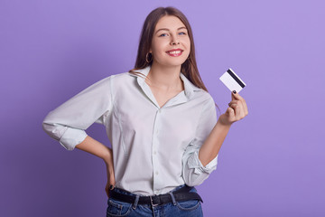 Portrait of funny attractive young female holding credit card in one hand, smiling sincerely, looking directly at camera, wearing jeans and light shirt, keeping hand on waist. Expenses concept.
