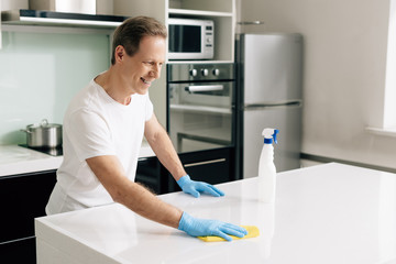 cheerful man in rubber gloves holding rag while cleaning kitchen table