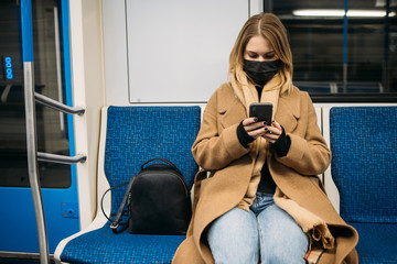 Blonde in medical mask with phone in her hands sitting in subway car.