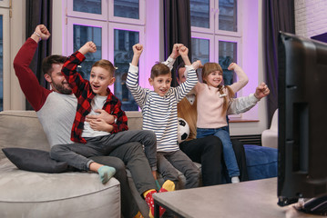 Happy bearded father and grandfather with three joyful grandchildren rejoicing the scored ball during watching football match on tv while spending time together at home.