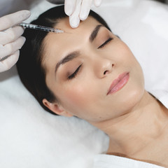 Calm peaceful young woman lying with closed eyes during botox injection in forehead area. Hand in white glove with syringe in hand doing beauty procedure for youthfull skin.