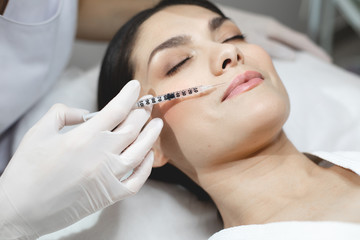 Calm peaceful young woman lying during botox injection process. Beautician in white gloves hold syringe at upper lip area. Doing injection careful and accurate.