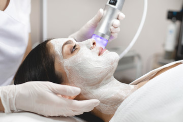 Obraz na płótnie Canvas Picture of laser grinding facial skin. Young woman lying with white cream mask upon her face. Skin treatment and spa at cosmetologist's room.